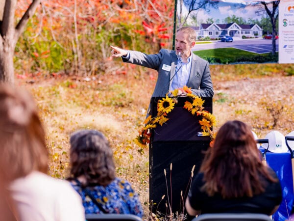 A white man wearing a blue blazer and light blue shirt standing at a podium set up outside points off into the distance.