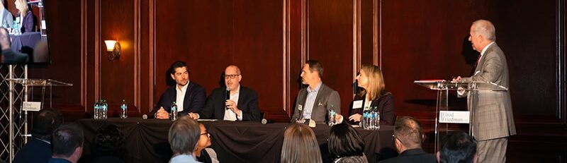 A group of people speak from a table in a front of others in a restaurant.