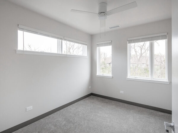 Bedroom with no furniture. Light streams through the windows and reflects on white walls and grey carpet.