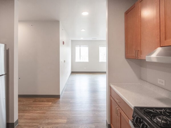Bright light streams into an empty apartment living room while looking from a kitchen with oak cabinets.