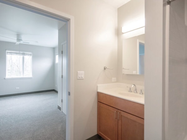 The view of an empty bedroom through the door of a bathroom. A sink is visible on the right side.