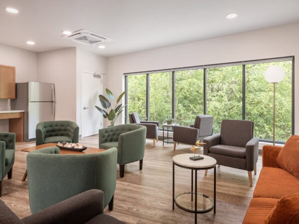 The interior of a community room with a wall of windows. Green trees visible through the windows. Chairs and side tables are arranged to encourage conversation.