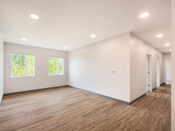 Interior of an empty apartment living room with white walls and greenery is visible through two large windows.