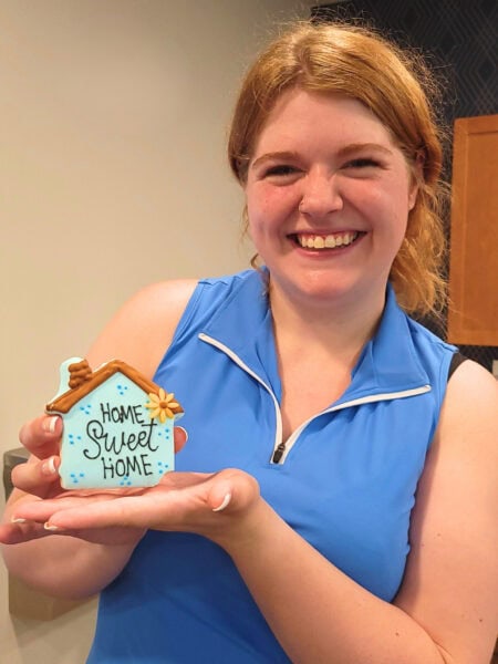 A woman with red hair, wearing a blue sleeveless dress, smiles while holding a cookie that says Home Sweet Home.