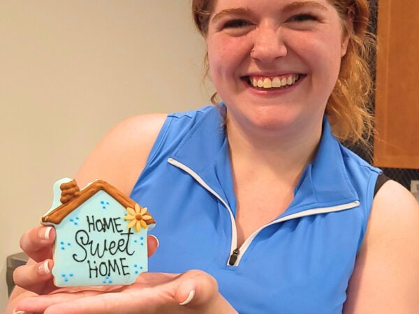 A woman with red hair, wearing a blue sleeveless dress, smiles while holding a cookie that says Home Sweet Home.