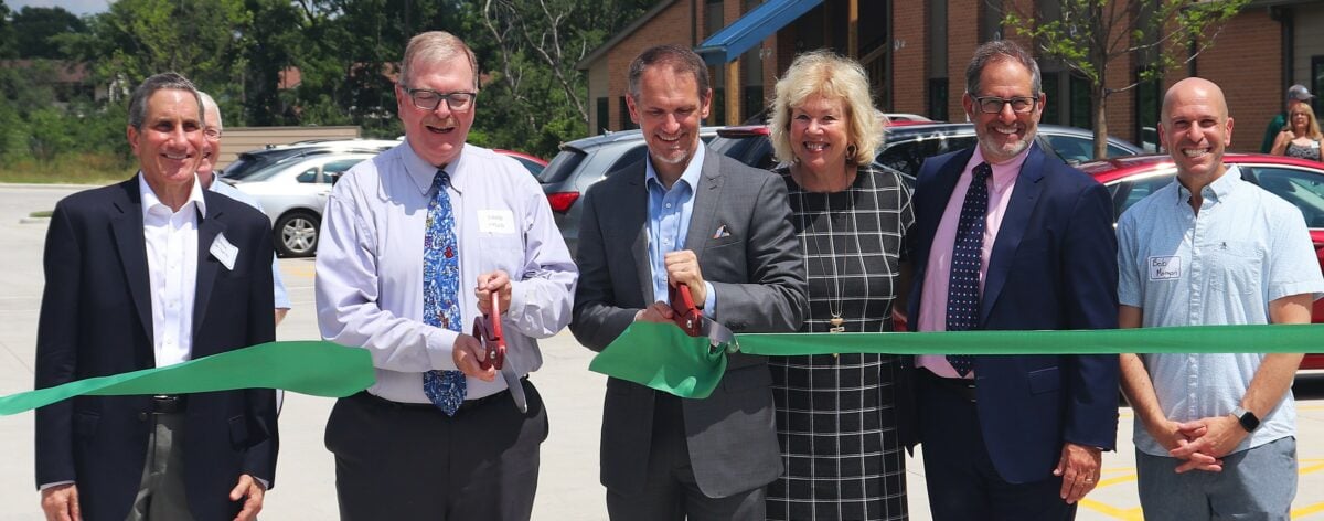 Professionally dressed men and women cut a large green ribbon in front of a two story apartment building.