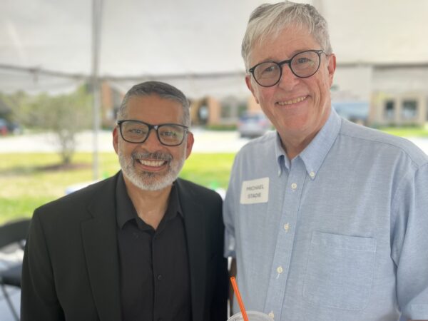 Two men pose together under a tent. The mood is festive and sunny.