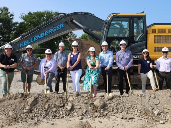 People wearing hard hats pose with shovels behind a mound of dirt and in front of a large construction vehicle.