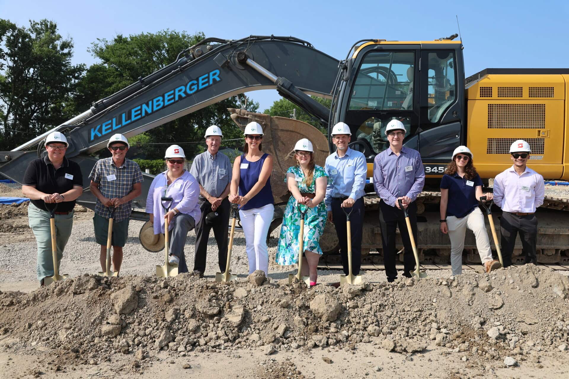 People wearing hard hats pose with shovels behind a mound of dirt and in front of a large construction vehicle.