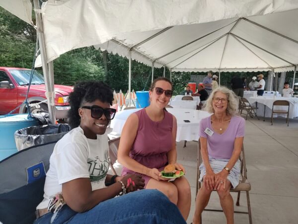 Three women pose sitting under a tent during a summer event.