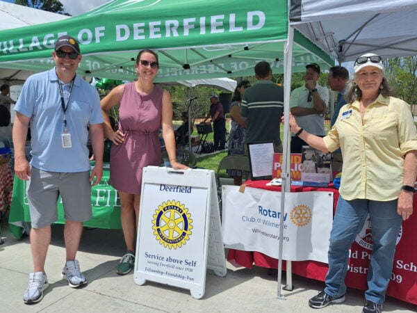 People pose in front of a tent during an outdoor event. The mood is friendly and feels like a warm summer day.
