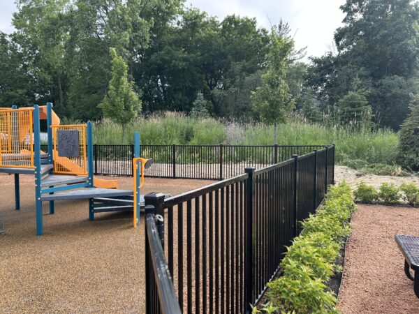 Green plants line a black metal fence that circles a children's playground.