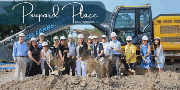 A group of people wearing hard hats toss dirt from shovels in front of a bulldozer.