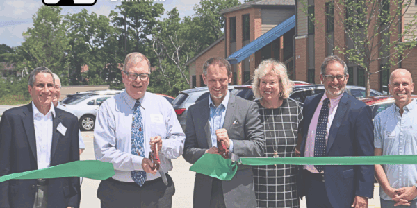 Professionally dressed men and women cut a large green ribbon in front of a two story apartment building.