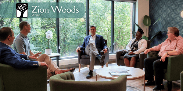 Professionally dressed people sit in armchairs in a community room with a wall of large windows. Trees and green leaves are visible through the windows.
