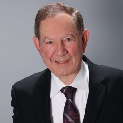 White man with brown/grey hair wearing a black suit and striped tie with a white shirt.