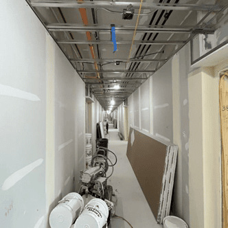 Long interior hallway with exposed wiring in the ceiling, walls partially mudded, and construction equipment lining the walls.