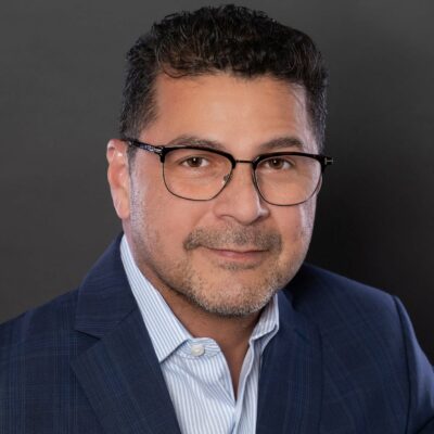 Man with brown short hair, wearing glasses and a navy blazer with a blue and white striped shirt.