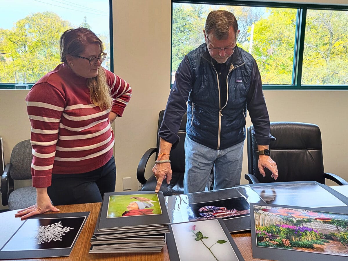 A woman in a red striped sweater and a man in a blue vest smile beside a table displaying colorful photographs. Large windows frame a lush green view outside.