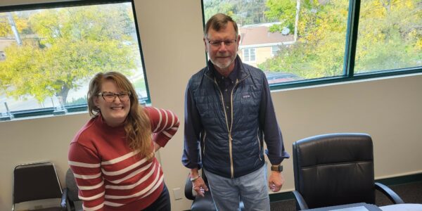 Two people review photographs on a table in a well-lit office. The woman wears a red striped sweater, and the man, a navy vest. The mood is focused.