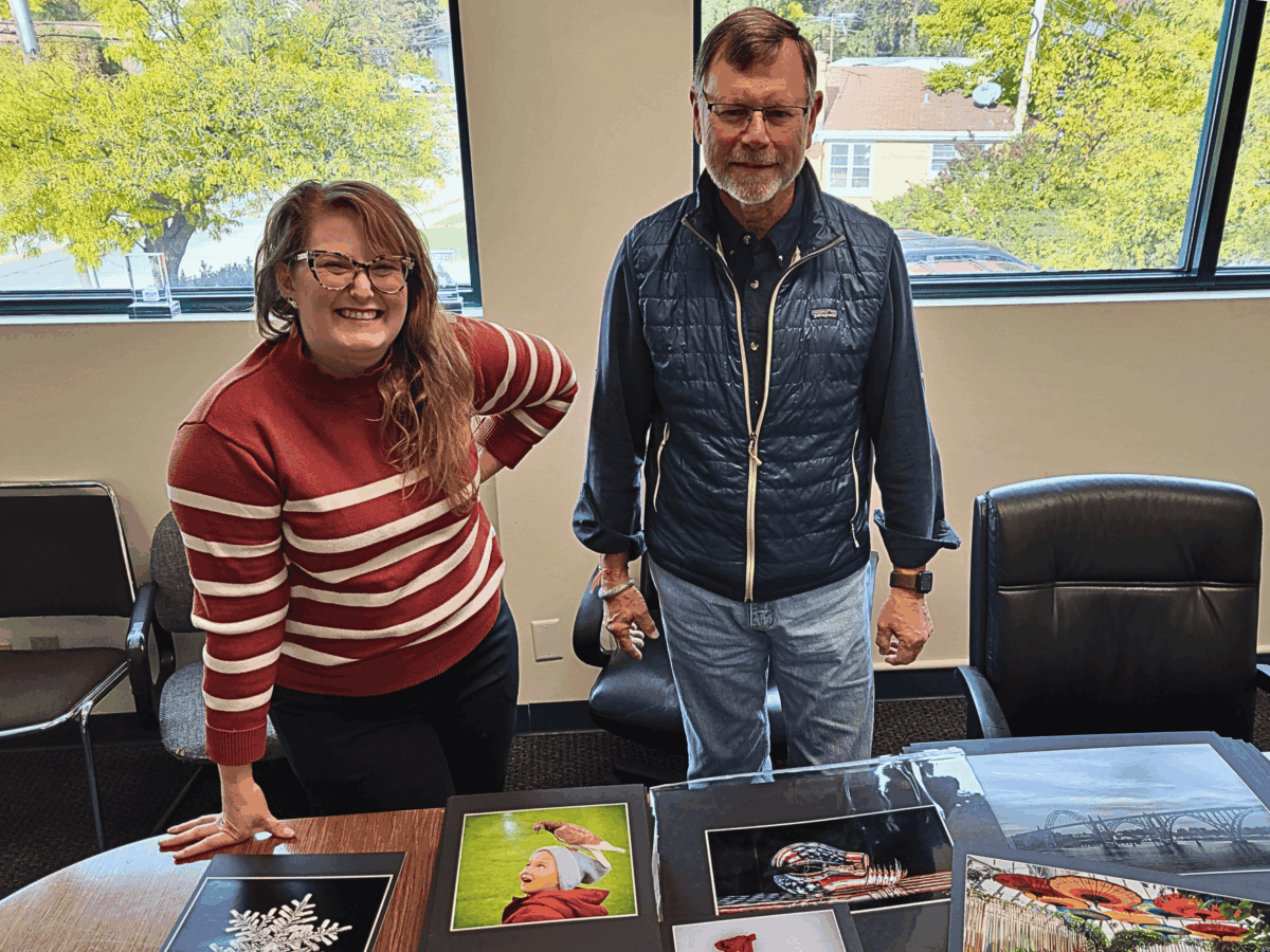 Two people review photographs on a table in a well-lit office. The woman wears a red striped sweater, and the man, a navy vest. The mood is focused.