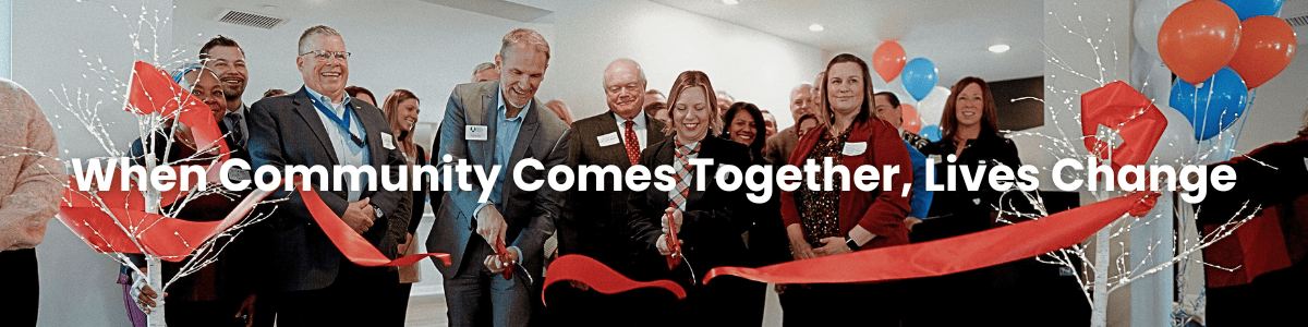 Professionally dressed people joyfully cut a wide red ribbon with oversized scissors. White text over the picture says When Community Comes Together, Lives Change.