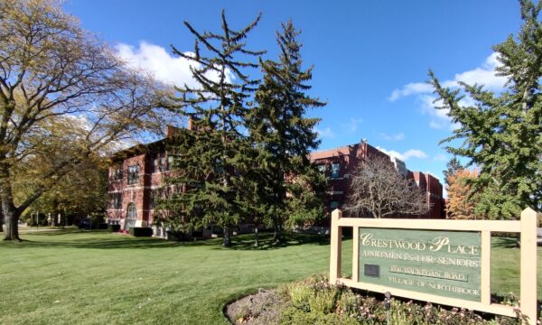 A red brick apartment building shaded by large trees with green leaves, a building sign in the foreground says Crestwood Place Apartments for Seniors. A blue sky in the background.