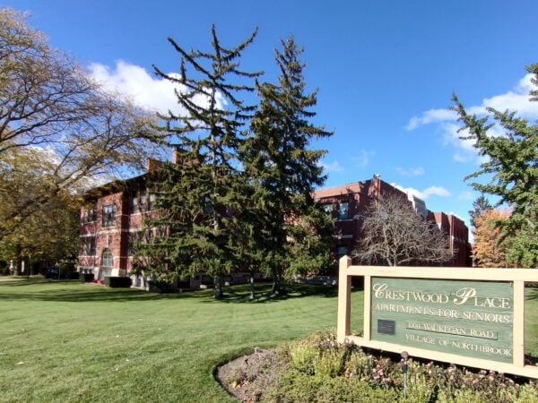 A red brick apartment building shaded by large trees with green leaves, a building sign in the foreground says Crestwood Place Apartments for Seniors. A blue sky in the background.