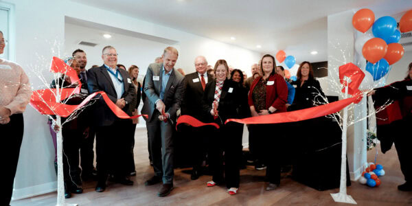 A group of people stand cheering as a man and woman cut a wide red ribbon using large scissions during a festive indoor ribbon-cutting ceremony. Bright balloons accentuate the joyful scene.