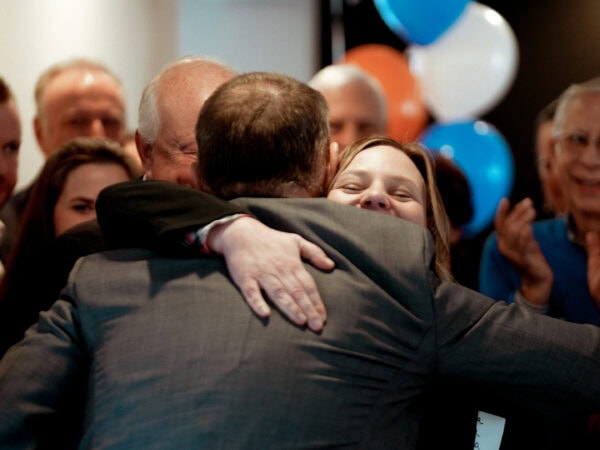 A joyful group celebrates with two people hugging warmly in the center. Smiling faces surround them, with colorful balloons in the background.