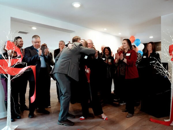 A group of people gathered in a room, celebrating a ribbon-cutting ceremony. Two individuals hug in the center, surrounded by others clapping. Festive balloons and decorations create a joyful atmosphere.