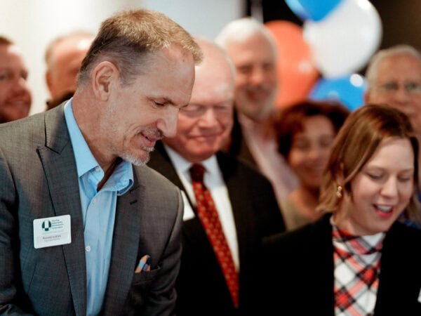A group of people is gathered at a cheerful event. A man and woman, both smiling, stand in the foreground. Blue, white, and orange balloons are visible in the background.