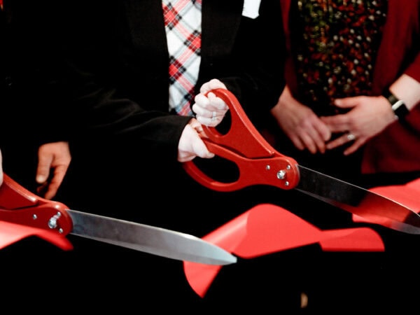Close-up of three people with large red scissors cutting a bright red ribbon at an event. The scene conveys a celebratory and formal tone.