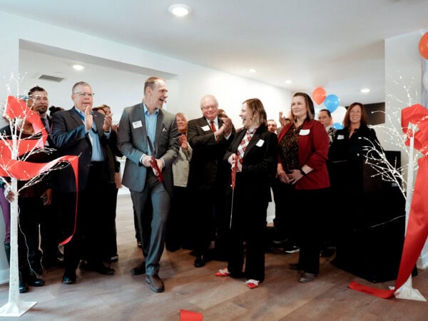 A group of people stand smiling at a ribbon-cutting ceremony inside a bright room. A man in a suit uses large scissors on the red ribbon. Bright balloons and decorative lights add a festive atmosphere.
