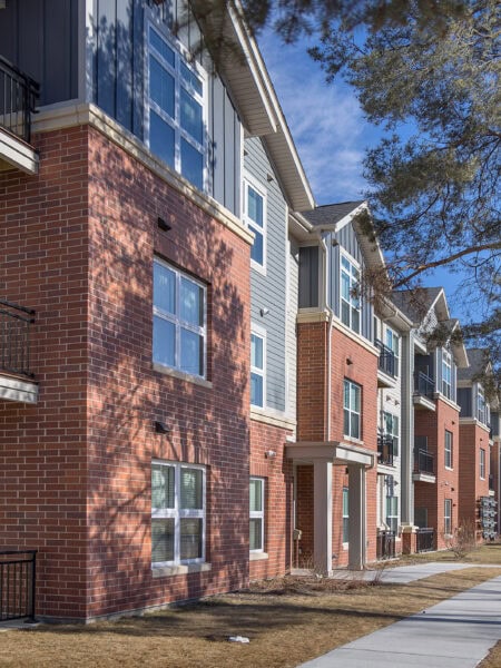A three-story apartment building with red brick and blue or tan siding. A sidewalk along the building is overhung with tree branches.