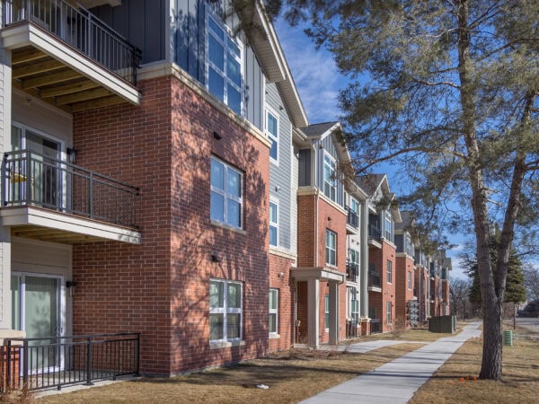 A three-story apartment building with red brick and blue or tan siding. A sidewalk along the building is overhung with tree branches.