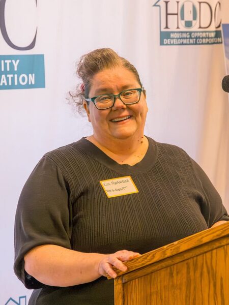 A woman with brown hair, wearing a dark shirt smiles behind a podium. A banner with the HODC logo is stretched behind her.