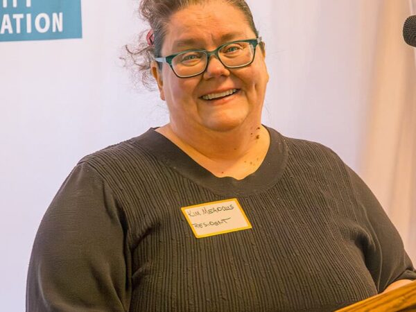 A woman with brown hair, wearing a dark shirt smiles behind a podium. A banner with the HODC logo is stretched behind her.