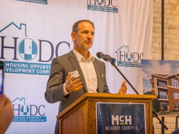 Man speaks into a microphone at a podium. HODC logo and apartment renderings behind him.