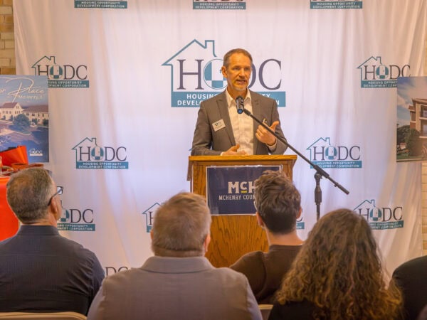 A man speaks at a podium labeled "McHenry County" to an audience, with HODC and housing development banners and architectural renderings displayed in the background.
