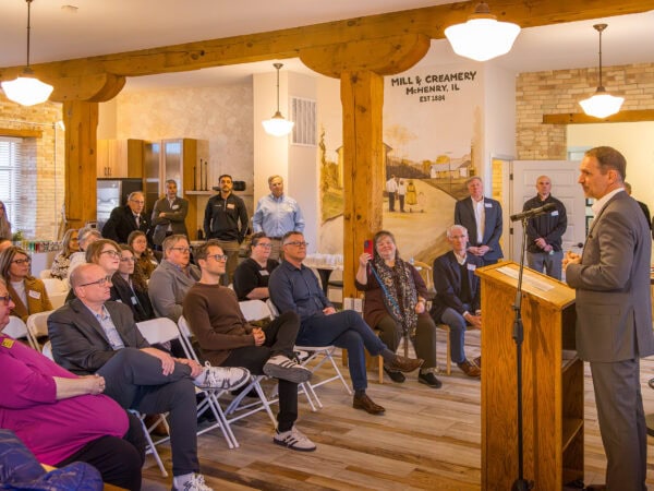 A man in a suit speaks at a podium to a seated audience in a room with wooden beams and a mural on the wall behind him.