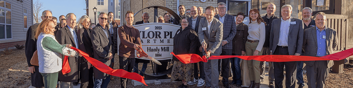 People posing with a red ribbon and large scissors, cutting the ribbon in front of the Taylor Place Apartments.