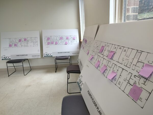 Four architectural floor plans are displayed on easels in a room. Each plan has purple sticky notes attached. Soft natural light enters through a window, creating a calm and organized atmosphere.