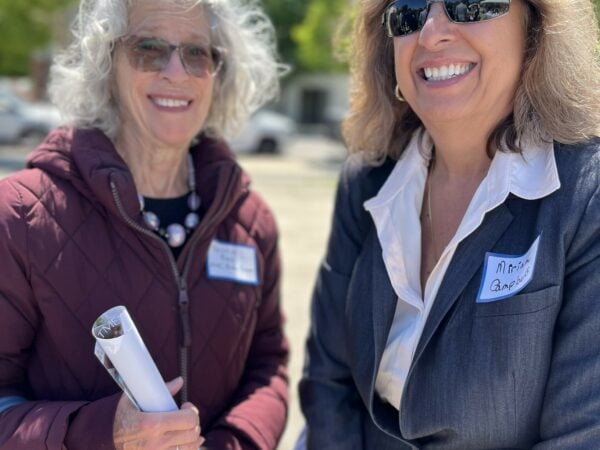 Two women wearing sunglasses pose together during an outdoor event.