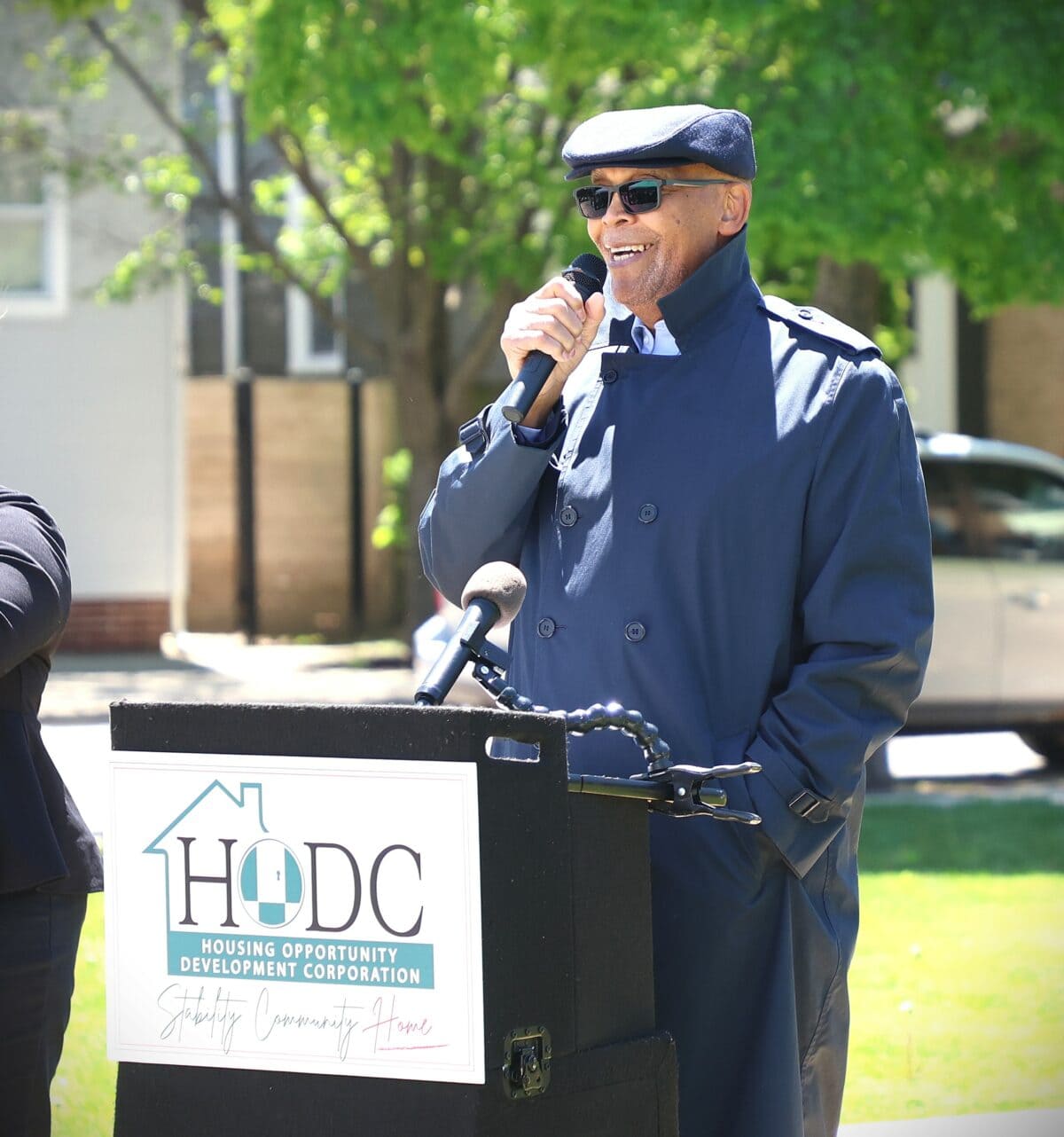 A man wearing a cap, sunglasses and a blue raincoat holds a microphone while speaking behind a podium labeled "HODC".