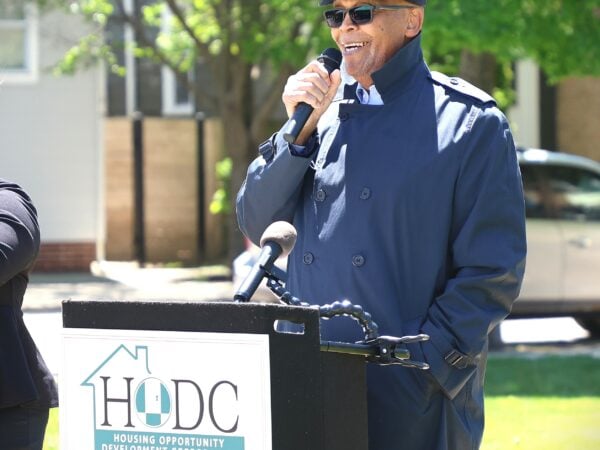 A man wearing a cap, sunglasses and a blue raincoat holds a microphone while speaking behind a podium labeled "HODC".