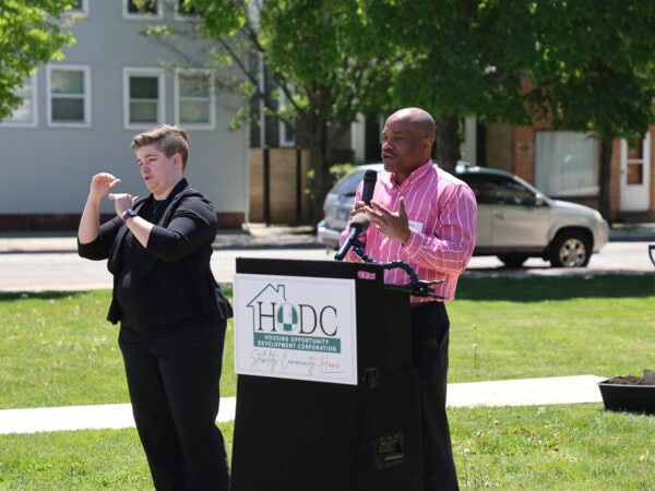A black man wearing a pink shirt speaks at a podium labeled "HODC" outdoors on a sunny day. A white woman with short hair and wearing a black outfit interprets in sign language. Green lawn and trees in the background.