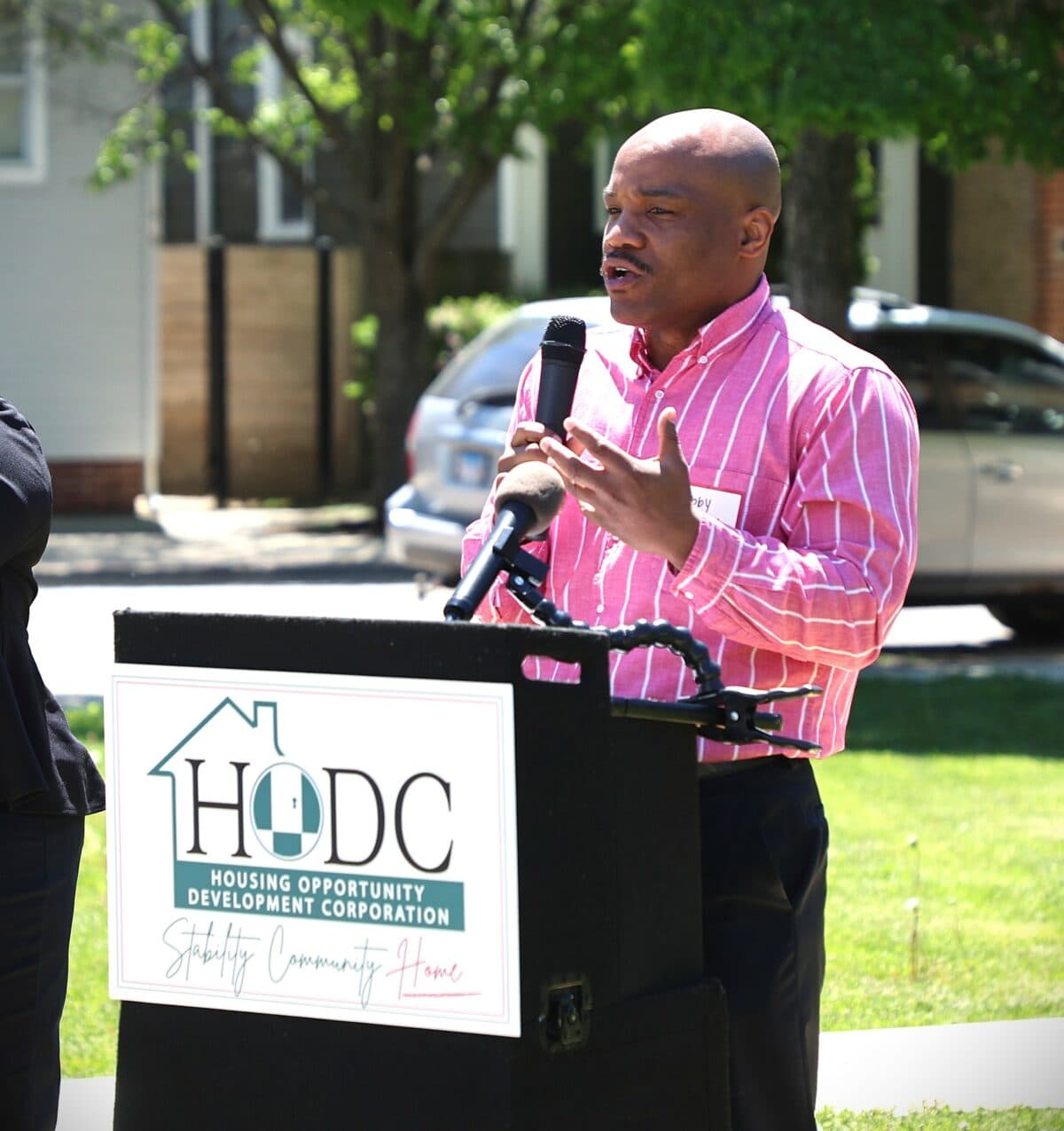 A black man wearing a pink shirt speaks at a podium labeled "HODC" outdoors on a sunny day. Green lawn and trees in the background.