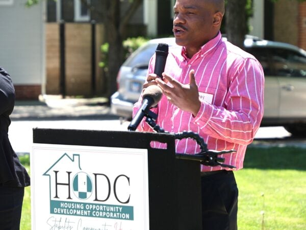 A black man wearing a pink shirt speaks at a podium labeled "HODC" outdoors on a sunny day. Green lawn and trees in the background.