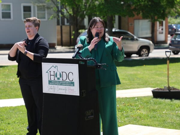 An Asian woman wearing green speaks at a podium labeled "HODC" outdoors on a sunny day, with white woman wearing black using sign language standing next to her. Green lawn and trees in the background.
